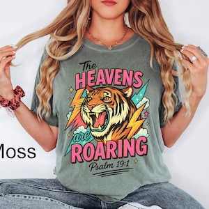 May include: Moss green t-shirt featuring a roaring tiger graphic with the text "The Heavens are Roaring" and "Psalm 19:1". The design includes pink, orange, yellow, and blue accents. The shirt is a relaxed fit.