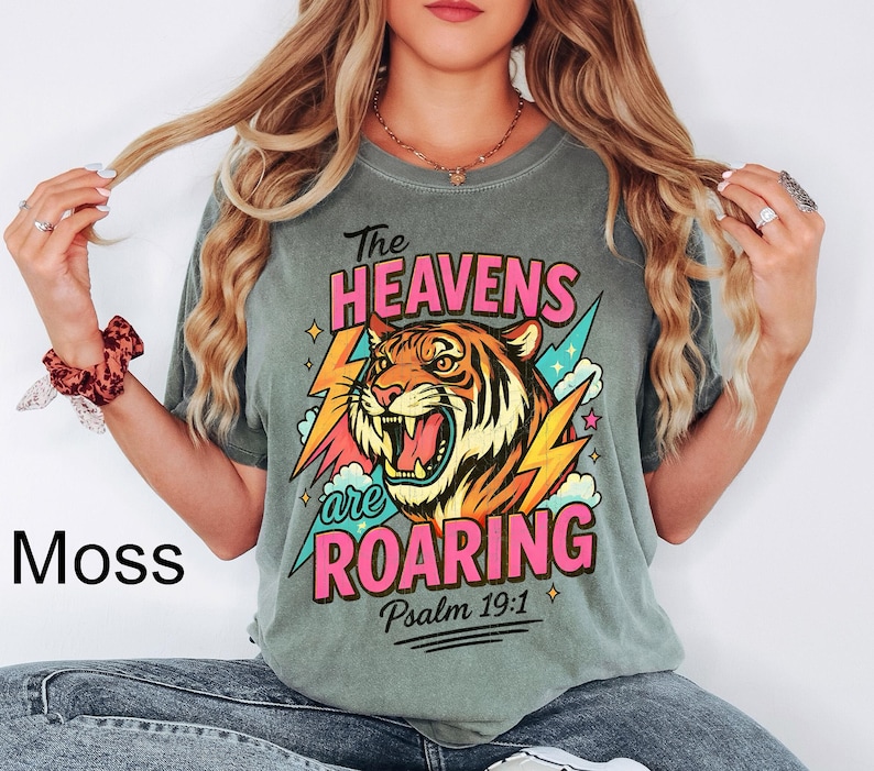 May include: Moss green t-shirt featuring a roaring tiger graphic with the text "The Heavens are Roaring" and "Psalm 19:1". The design includes pink, orange, yellow, and blue accents. The shirt is a relaxed fit.