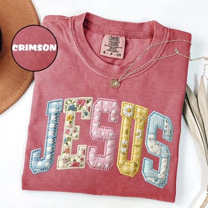 May include: A crimson short-sleeved t-shirt featuring the word JESUS in large, fabric-appliqued varsity-style letters. Each letter displays a different pattern, including blue gingham, floral prints, pink polka dots, and yellow gingham.