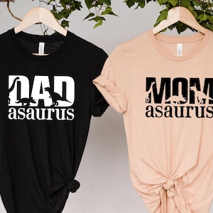 May include: Two t-shirts, one black and one peach, hanging on wooden hangers. The black shirt has the text "DAD asaurus" in white, with dinosaur silhouettes. The peach shirt says "MOM asaurus" in black, also with dinosaur silhouettes.