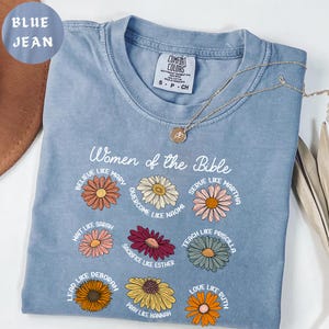 May include: A blue jean colored t-shirt with the text "Women of the Bible" and floral illustrations. The shirt features the words "Believe like Mary", "Serve like Martha", and other inspirational phrases. A gold necklace is draped across the shirt.