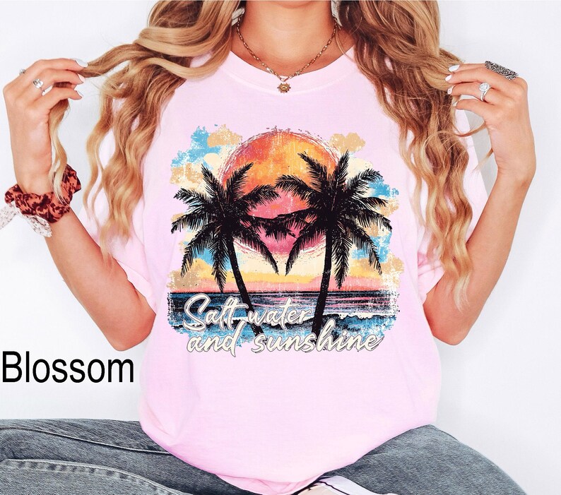 May include: A light pink t-shirt featuring a sunset scene with palm trees and the text "Salt water and sunshine." The graphic includes orange, yellow, blue, and black colors. The shirt is a casual wear item.