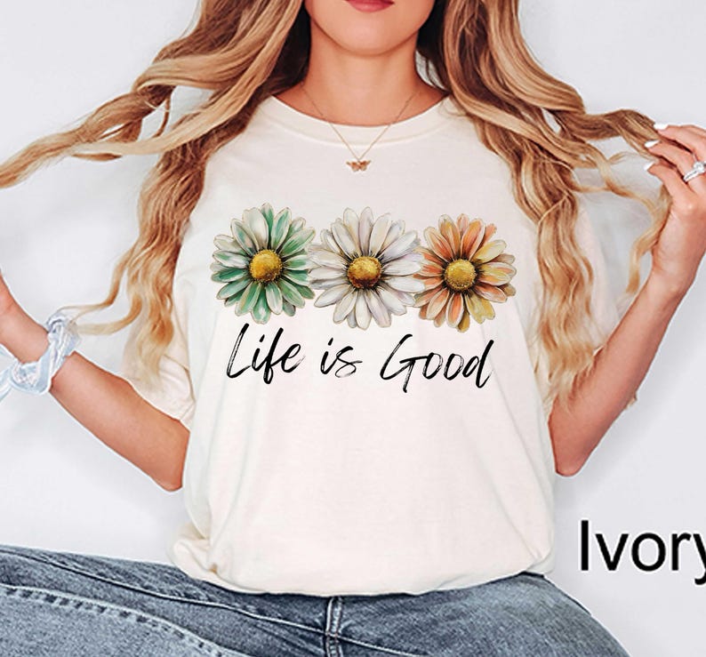 May include: Ivory-colored t-shirt featuring three daisy flowers in green, white, and orange with yellow centers. The phrase "Life is Good" is written in black script below the flowers. The shirt is a casual, everyday wear item.