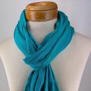 May include: A teal-colored scarf draped around a mannequin's neck. The scarf is made of a textured fabric and is knotted at the front. The mannequin has a wooden neck piece. The background is a plain white wall.