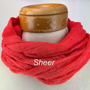 Puede incluir: Una bufanda roja transparente drapeada alrededor del cuello de un maniquí. La bufanda está hecha de una tela ligera y fluida, con una textura suave y drapeada. La palabra "Sheer" está impresa en la bufanda.