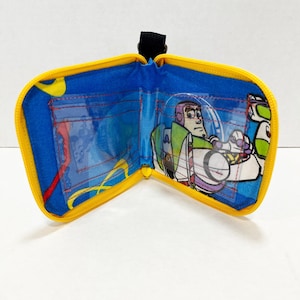 May include: Open blue and yellow zippered wallet featuring a cartoon image of Buzz Lightyear. The wallet has a black strap and clear pockets. The interior is blue with yellow accents and red stitching.