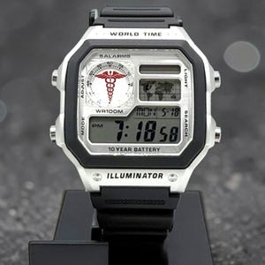 May include: A silver and black digital watch with the text "WORLD TIME" and "DALARMS". The watch face displays the time, date, and a medical symbol. The watch is on a black stand. The watch has a black band and the text "10 YEAR BATTERY".