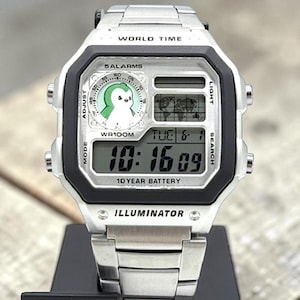 May include: A silver digital wristwatch with a black bezel and a stainless steel band. The watch face displays the time, date, and a penguin graphic. The text "WORLD TIME", "5 ALARMS", "WR100M", "10 YEAR BATTERY", and "ILLUMINATOR" are visible.
