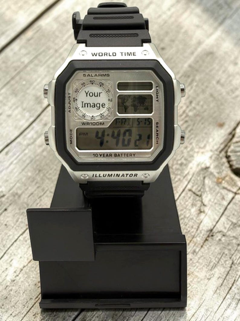 Pode incluir: Um rel&oacute;gio de pulso digital com uma pulseira preta e um mostrador quadrado prateado. O rel&oacute;gio exibe a hora, a data e outras fun&ccedil;&otilde;es. O mostrador inclui o texto "WORLD TIME", "5 ALARMS", "WR100M", "10 YEAR BATTERY" e "ILLUMINATOR". O rel&oacute;gio est&aacute; em um suporte preto.