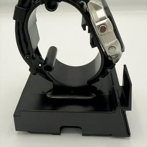 May include: A black watch display stand holding a silver and black wristwatch. The watch has a black band and a silver face with red accents. The stand is made of black plastic and has a curved design to hold the watch.