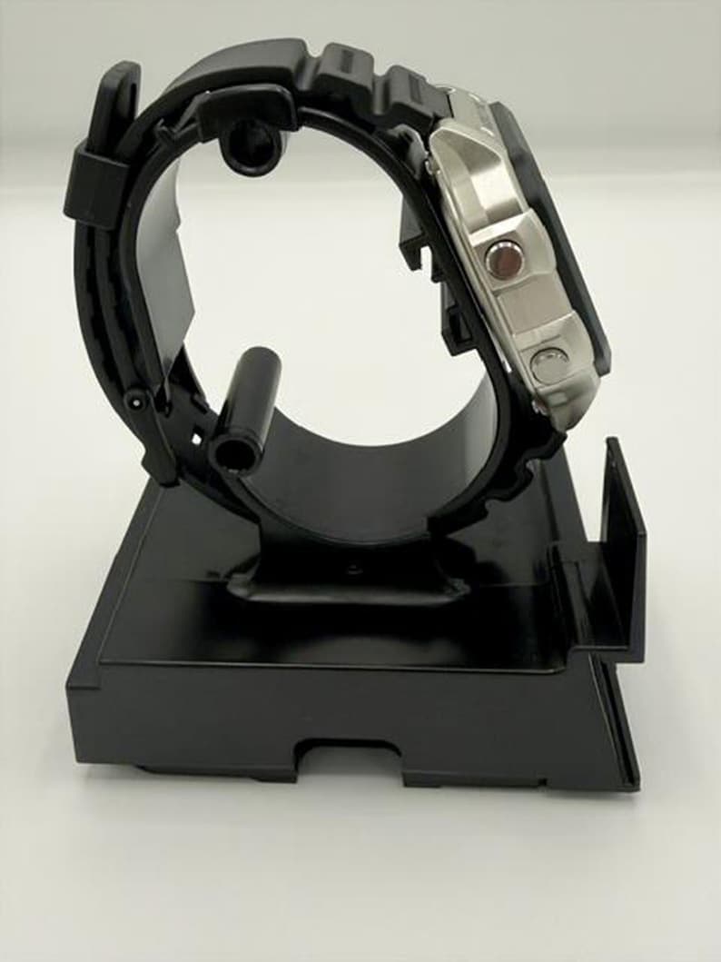 May include: A black watch display stand holding a silver and black wristwatch. The watch has a black band and a silver face with red accents. The stand is made of black plastic and has a curved design to hold the watch.