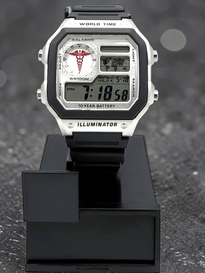 May include: A silver and black digital watch with the text "WORLD TIME" and "DALARMS". The watch face displays the time, date, and a medical symbol. The watch is on a black stand. The watch has a black band and the text "10 YEAR BATTERY".