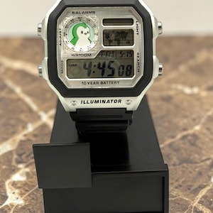 May include: A digital wristwatch with a black strap and a silver-colored face. The watch displays the time, date, and a world map. The face features a penguin graphic and the text "WORLD TIME", "SALARMS", and "ILLUMINATOR".
