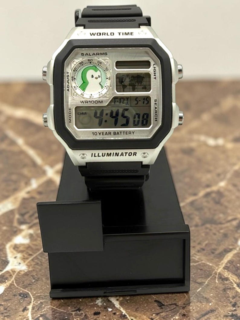 May include: A digital wristwatch with a black strap and a silver-colored face. The watch displays the time, date, and a world map. The face features a penguin graphic and the text "WORLD TIME", "SALARMS", and "ILLUMINATOR".