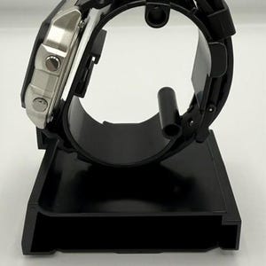 Pode incluir: Um rel&oacute;gio digital preto com mostrador prateado e pulseira de borracha preta &eacute; exibido em um suporte de rel&oacute;gio preto. O rel&oacute;gio est&aacute; inclinado para mostrar o perfil lateral, destacando os bot&otilde;es e os detalhes do mostrador.