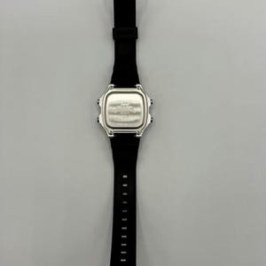 Pode incluir: Um rel&oacute;gio digital preto com mostrador prateado e pulseira preta. O rel&oacute;gio tem um mostrador retangular com cantos arredondados e uma fivela preta. A pulseira tem v&aacute;rios orif&iacute;cios para ajuste.