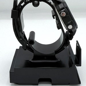 May include: A black digital watch with a black band is displayed on a black stand. The watch has a square face with two silver buttons on the side. The band is made of a textured material.