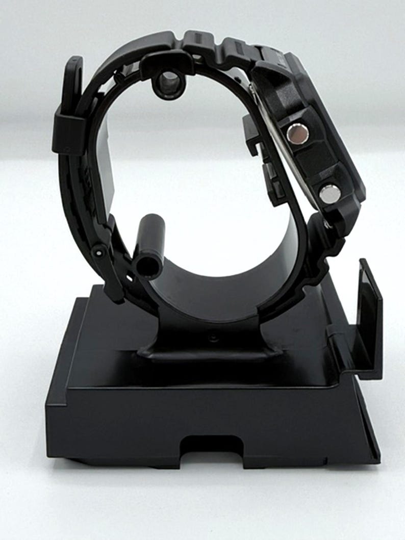 May include: A black digital watch with a black band is displayed on a black stand. The watch has a square face with two silver buttons on the side. The band is made of a textured material.