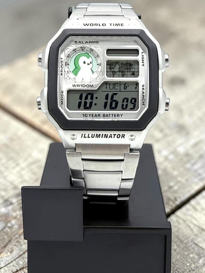 May include: A silver digital wristwatch with a black bezel and a stainless steel band. The watch face displays the time, date, and a penguin graphic. The text "WORLD TIME", "5 ALARMS", "WR100M", "10 YEAR BATTERY", and "ILLUMINATOR" are visible.