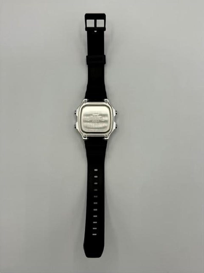 May include: A black digital watch with a silver-colored face and a black band. The watch has a rectangular face with rounded corners and a black buckle. The band has multiple holes for adjustment.