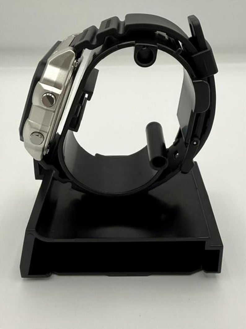 May include: A black digital watch with a silver-tone face and a black rubber strap is displayed on a black watch stand. The watch is angled to show the side profile, highlighting the buttons and details of the watch face.