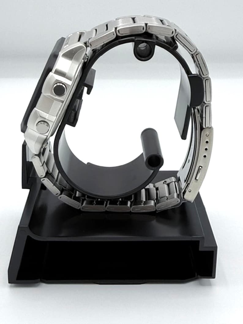 May include: A silver-toned wristwatch with a stainless steel band is displayed on a black watch stand. The watch face is rectangular with two circular buttons. The band is composed of small, rectangular links.