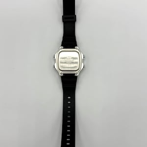 May include: A black digital watch with a silver face and a black rubber strap. The watch has a rectangular face and a buckle closure. The watch is shown against a white background.