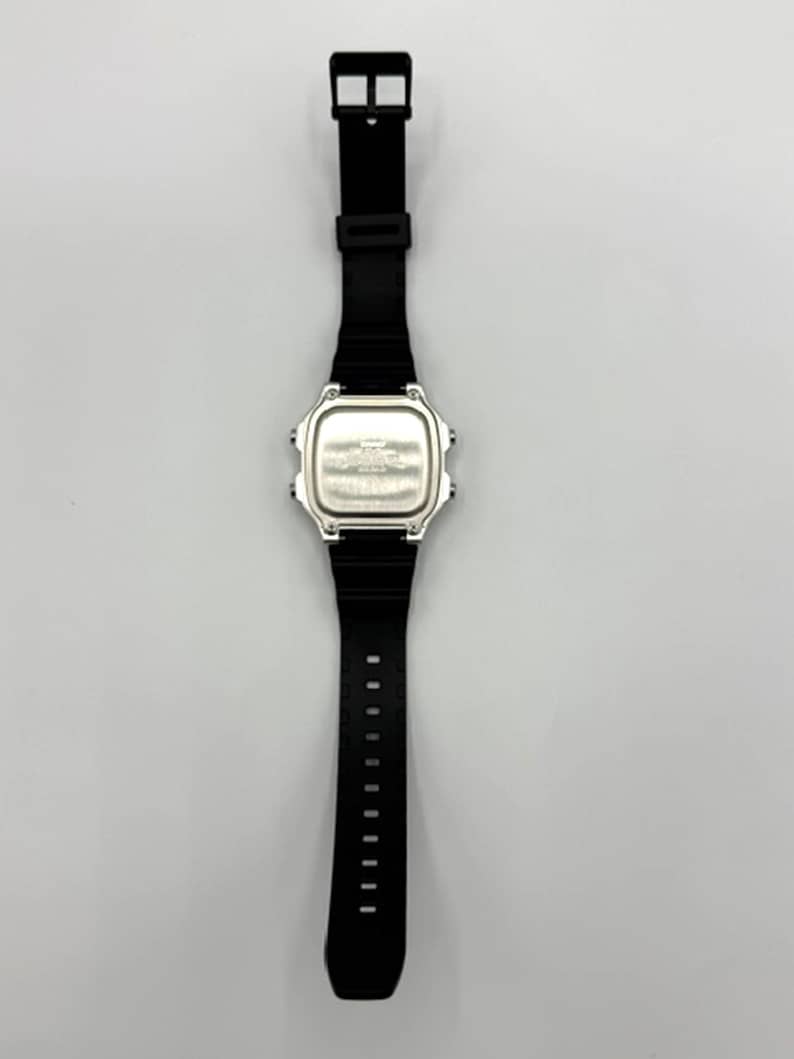 May include: A black digital watch with a silver face and a black rubber strap. The watch has a rectangular face and a buckle closure. The watch is shown against a white background.