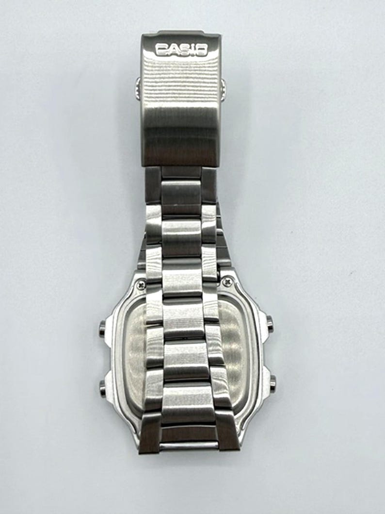 May include: A stainless steel Casio digital watch, viewed from the back. The watch features a rectangular face with a silver-tone metal band and clasp. The word "CASIO" is embossed on the clasp.