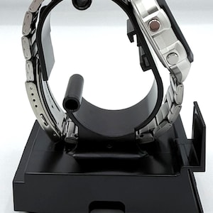 May include: A silver-toned wristwatch with a stainless steel band is displayed on a black stand. The watch face is square with a digital display. The band is composed of small, rectangular links. The watch is presented on a black, rectangular stand.