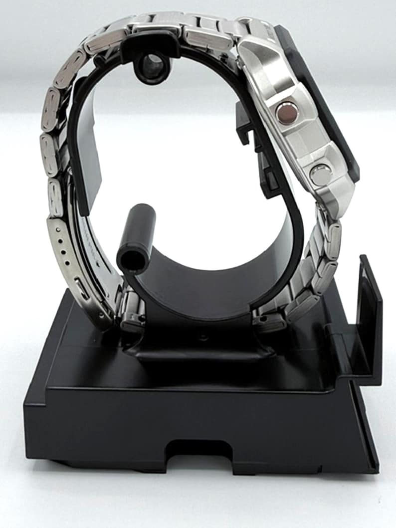 May include: A silver-toned wristwatch with a stainless steel band is displayed on a black stand. The watch face is square with a digital display. The band is composed of small, rectangular links. The watch is presented on a black, rectangular stand.