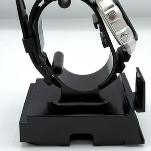 May include: A black wristwatch with a silver face is displayed on a black watch stand. The watch has a black rubber strap and a square silver case. The stand is rectangular with a curved top to hold the watch.