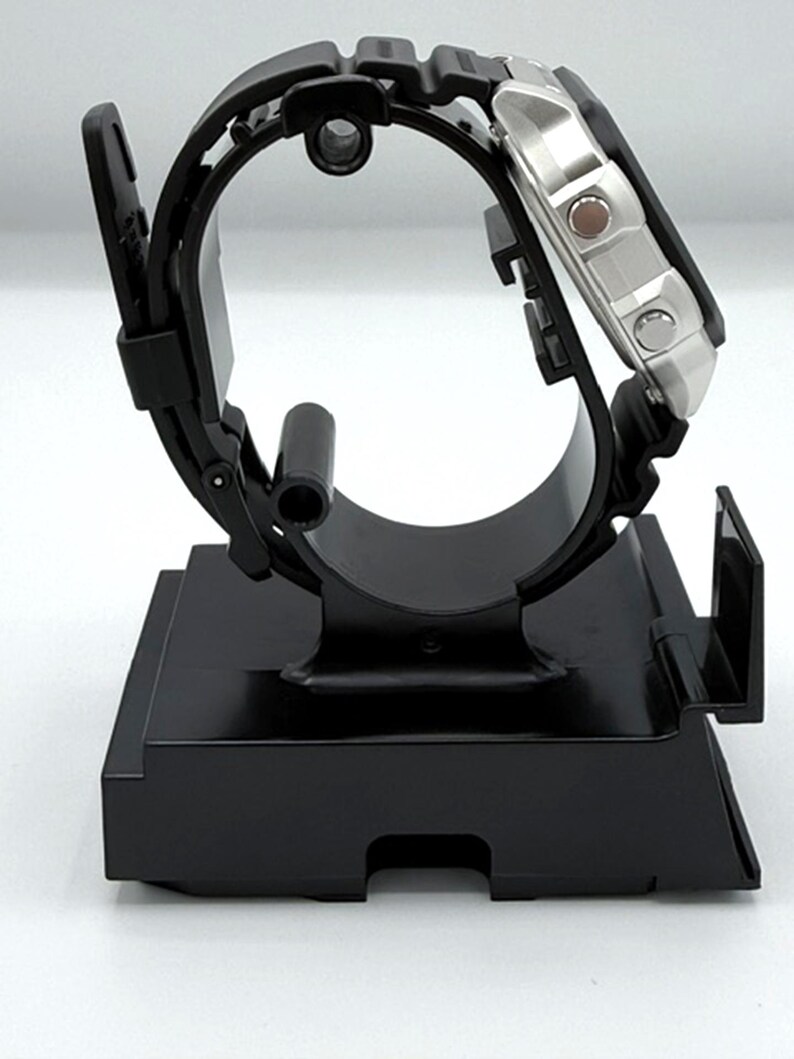 May include: A black wristwatch with a silver face is displayed on a black watch stand. The watch has a black rubber strap and a square silver case. The stand is rectangular with a curved top to hold the watch.