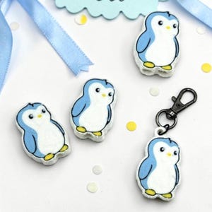 May include: Three small penguin-shaped charms and one penguin-shaped keychain with a black metal lobster clasp. Each penguin figure features a white belly, light blue wings and head, and yellow feet, rendered in a flat, cartoon-style design.