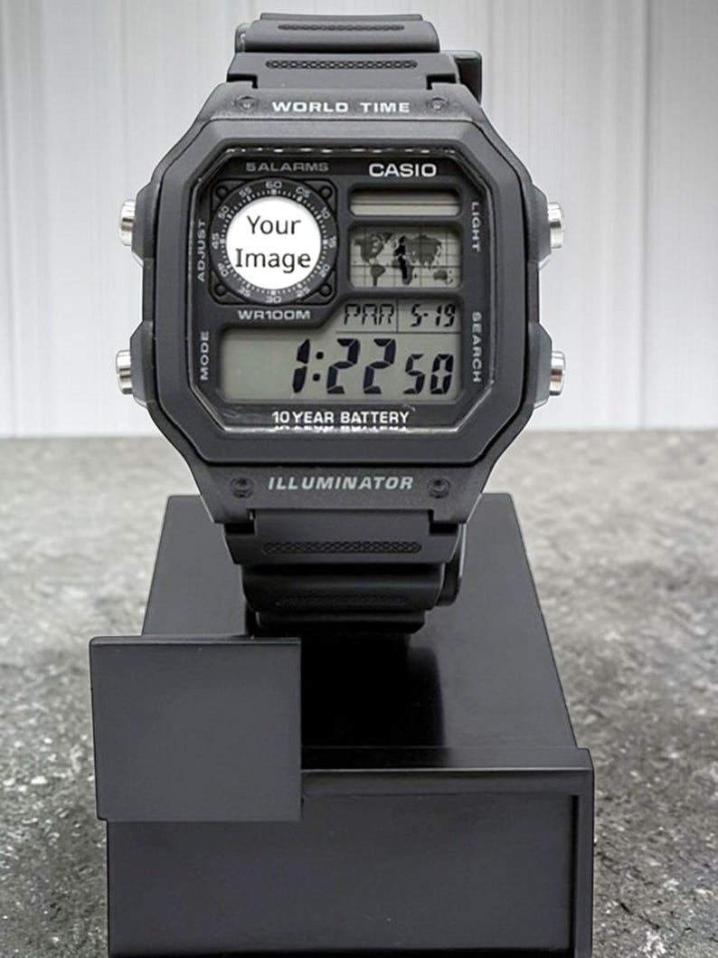 May include: Black Casio digital watch with a rectangular face, displaying the time and date. The watch features multiple buttons and a black wristband. The watch is on a black display stand.