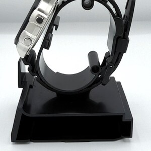 May include: A black digital watch with a silver face and black rubber strap is displayed on a black watch stand. The watch is angled, showcasing the face and side profile. The stand has a rectangular base and a curved support.