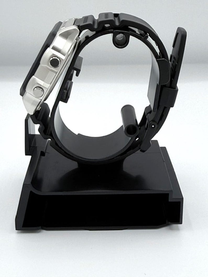 May include: A black digital watch with a silver face and black rubber strap is displayed on a black watch stand. The watch is angled, showcasing the face and side profile. The stand has a rectangular base and a curved support.
