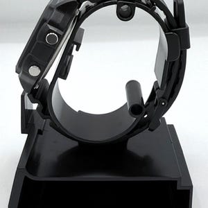 May include: A black digital watch with a black band is displayed on a black watch stand. The watch has a square face with buttons on the side. The stand is designed to hold the watch securely, showcasing its design.