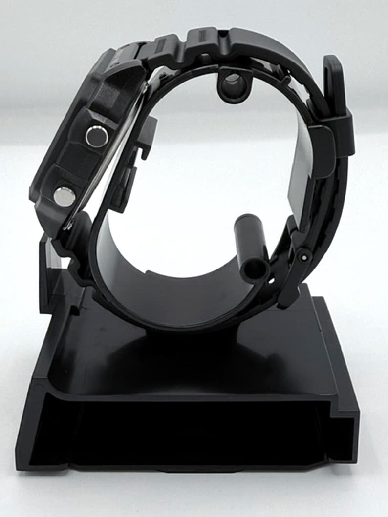 May include: A black digital watch with a black band is displayed on a black watch stand. The watch has a square face with buttons on the side. The stand is designed to hold the watch securely, showcasing its design.