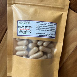 May include: A brown paper bag with a clear window displaying numerous beige capsules. The bag features a purple label with the text "MSM with whole food Vitamin C" and additional text detailing ingredients and usage. The bag is sealed and sits on a wooden surface.