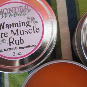 Puede incluir: Dos latas plateadas con etiquetas rosas. La etiqueta de la lata de la izquierda dice "Wonder Weeds Warming Sore Muscle Rub All Natural Ingredients 2 oz.". La lata de la derecha es parcialmente visible y tiene una etiqueta rosa con la palabra "Sore" visible.