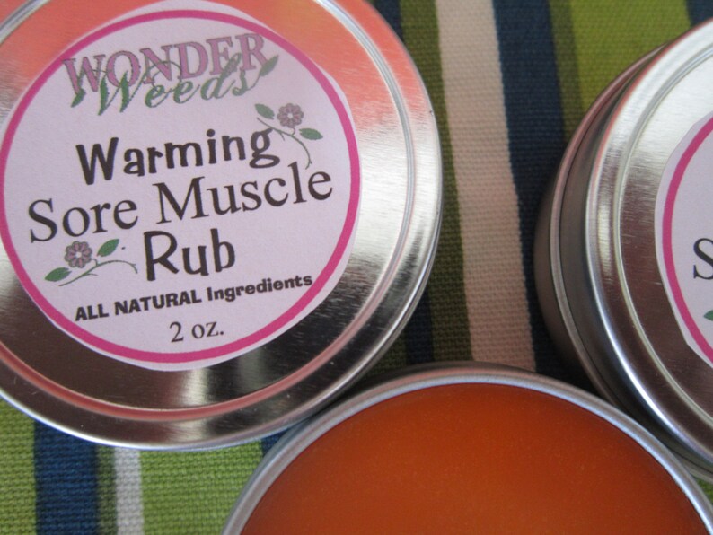 Puede incluir: Dos latas plateadas con etiquetas rosas. La etiqueta de la lata de la izquierda dice "Wonder Weeds Warming Sore Muscle Rub All Natural Ingredients 2 oz.". La lata de la derecha es parcialmente visible y tiene una etiqueta rosa con la palabra "Sore" visible.
