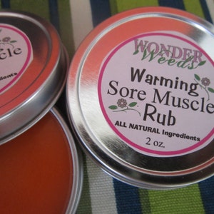 Puede incluir: Dos latas plateadas con etiquetas rosas. La etiqueta de la lata superior dice "Warming Sore Muscle Rub" con el texto "Wonder Weeds" encima. La etiqueta de la lata inferior dice "Warming Muscle Rub". Ambas etiquetas tienen el texto "All Natural Ingredients" y "2 oz."