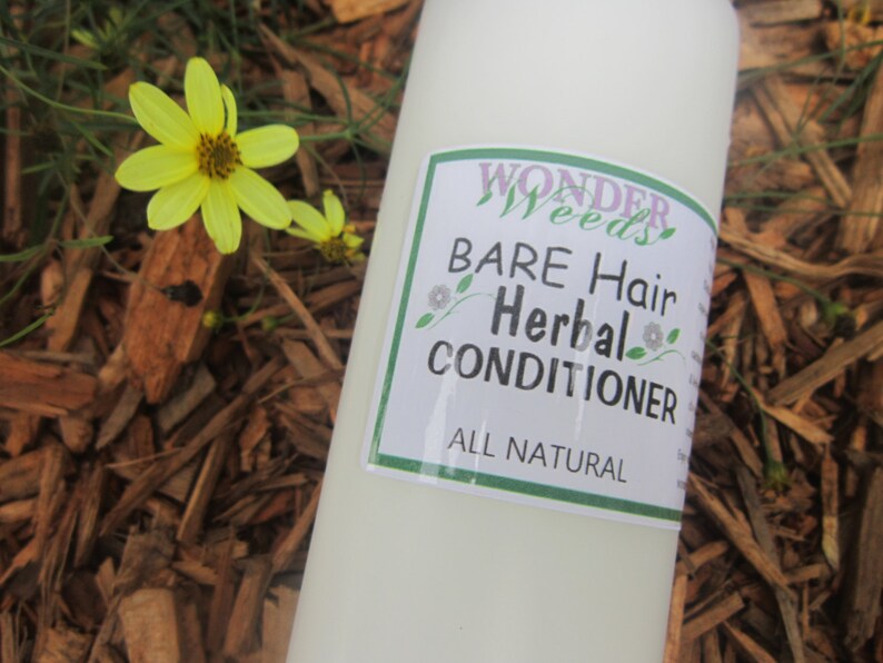 May include: A white bottle of "Wonder Weeds" Bare Hair Herbal Conditioner with a green and white label. The bottle is next to yellow flowers and wood chips.