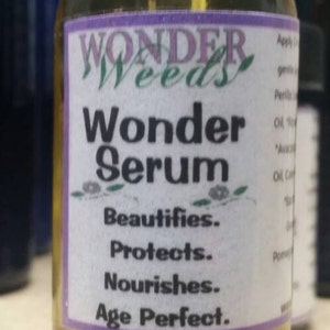 May include: A small glass bottle with a yellow liquid inside. The label is white with purple trim and reads "WONDER Weeds Wonder Serum Beautifies. Protects. Nourishes. Age Perfect."