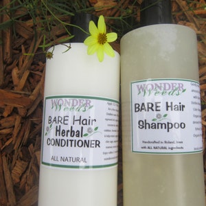 May include: Two bottles of Wonder Weeds Bare Hair Herbal Conditioner and Shampoo. The bottles are white with green and purple labels. A yellow flower sits atop one bottle. The background is brown wood chips.