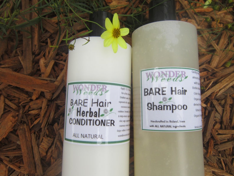 May include: Two bottles of Wonder Weeds Bare Hair Herbal Conditioner and Shampoo. The bottles are white with green and purple labels. A yellow flower sits atop one bottle. The background is brown wood chips.