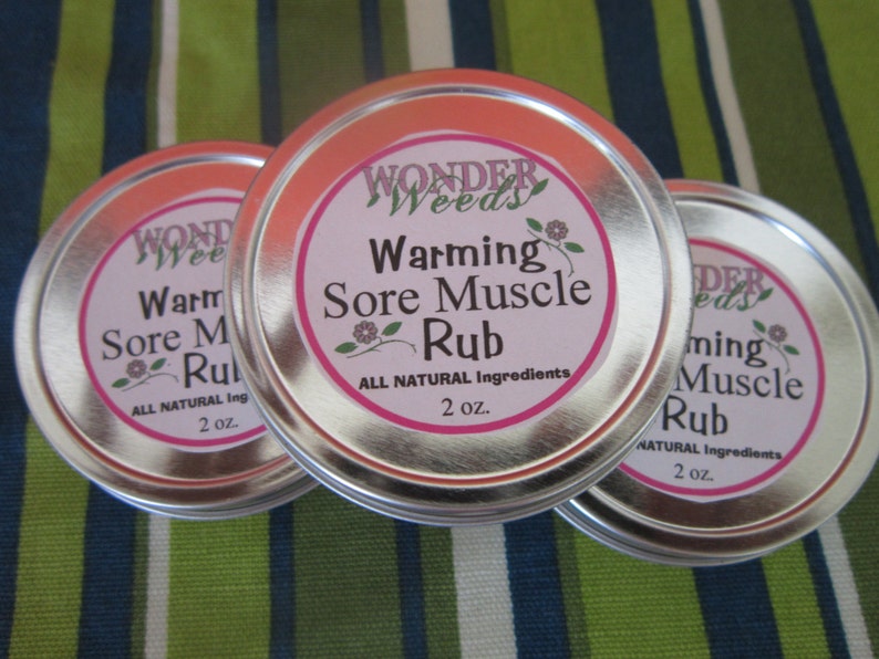 Puede incluir: Tres latas plateadas con etiquetas blancas y letras rosas. Las etiquetas dicen "Wonder Weeds Warming Sore Muscle Rub" y "All Natural Ingredients 2 oz."