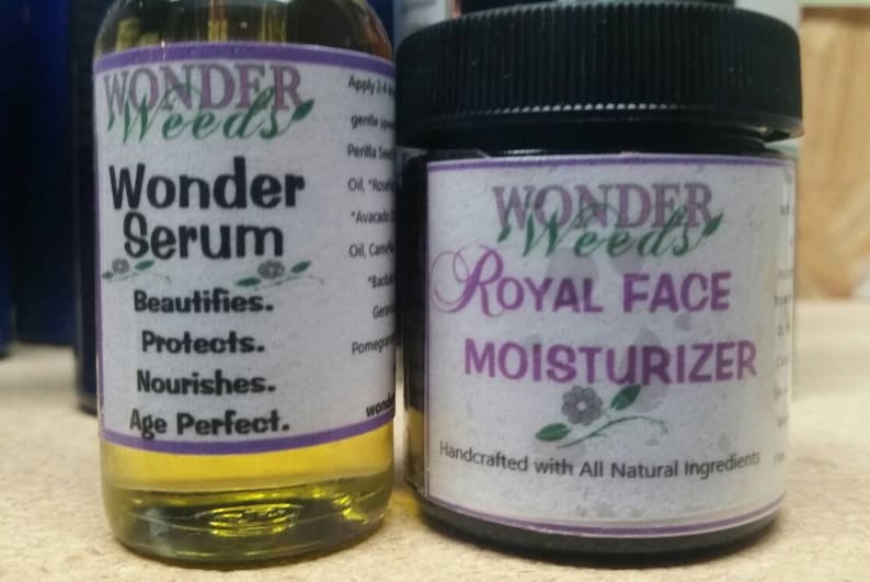 May include: Two bottles of skin care products. The bottle on the left is a clear glass bottle with a yellow liquid and a purple label that says "Wonder Weeds Wonder Serum Beautifies. Protects. Nourishes. Age Perfect." The bottle on the right is a dark glass jar with a black lid and a purple label that says "Wonder Weeds Royal Face Moisturizer Handcrafted with All Natural Ingredients."