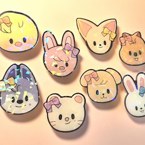 May include: A collection of eight holographic stickers featuring adorable animal faces, including a chick, fox, pig, dog, cat, and rabbits. Each sticker has a bow and a black outline. The stickers are in various colors, including yellow, pink, and white.